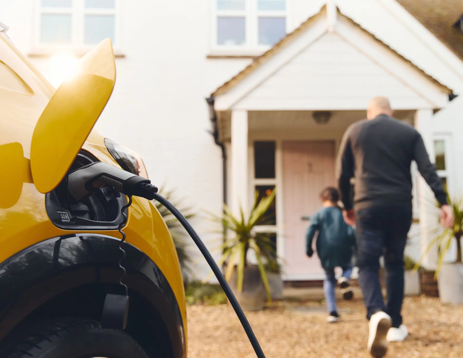 A yellow electric vehicle being charged with a home EV charger outside a house, a man and his young child are walking to the front door 